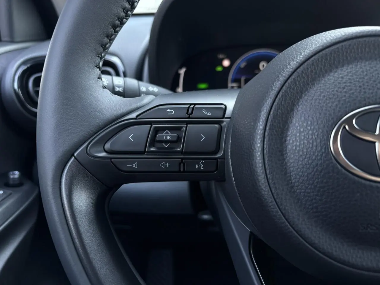 Gros plan sur le côté gauche du volant cuir noir de la Toyota Yaris 2025, avec commandes intégrées et tableau de bord numérique en arrière-plan.