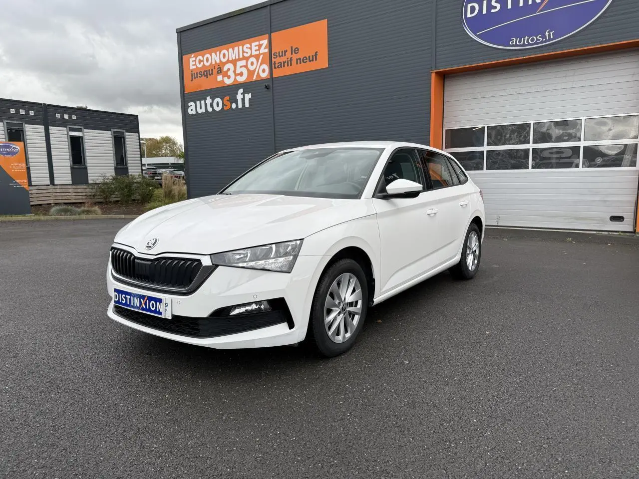 Vue 3/4 avant d'une Skoda Scala blanche stationnée devant un bâtiment commercial avec jantes alliage argent.