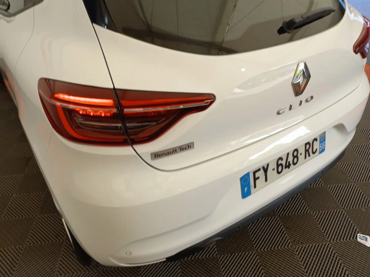Vue 3/4 arrière droite d'une Renault Clio blanche avec feux LED allumés et badge Renault Tech visible sur le coffre.