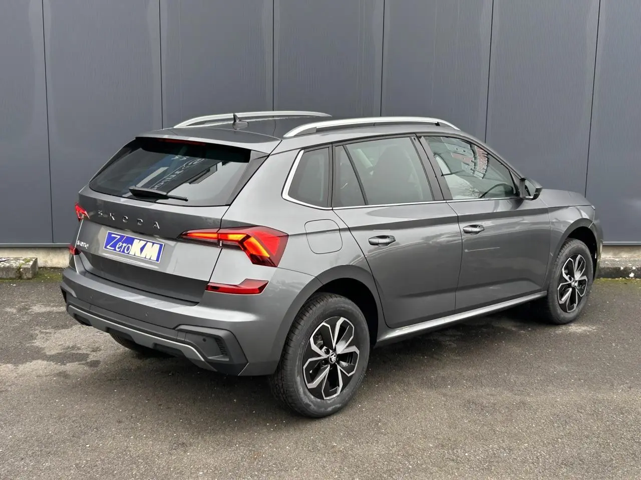 Vue 3/4 arrière droite d'un Skoda Kamiq gris graphite avec vitres surteintées et barres de toit argentées.
