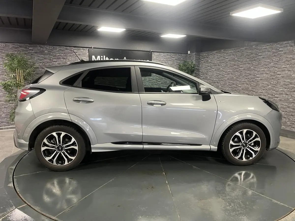 Ford Puma gris Solar en profil droit, avec jantes alliage noires et barres de toit noires visibles en intérieur showroom.