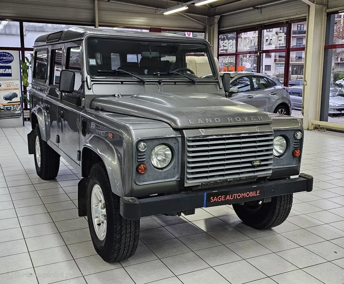 Vue 3/4 avant d'un Land Rover Defender 110 Station Wagon S gris, avec calandre distinctive et phare rond en intérieur showroom.