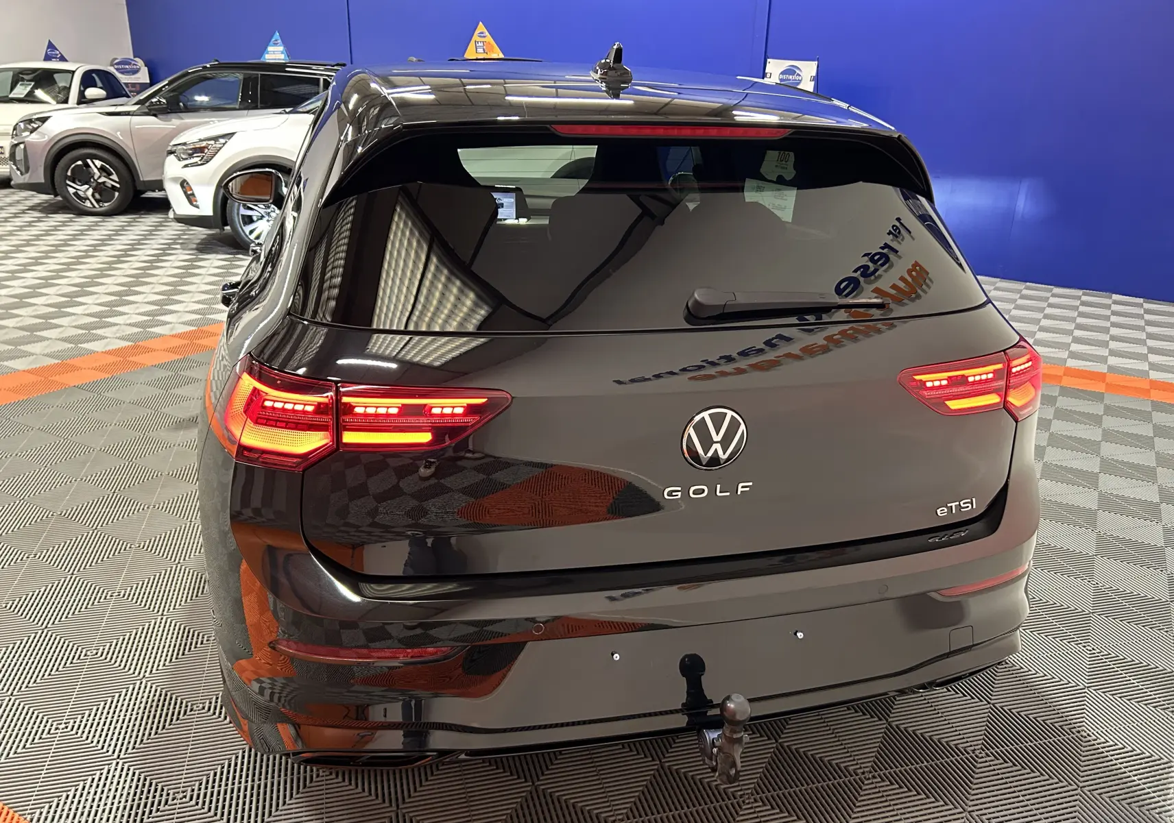 Vue arrière d'une Volkswagen Golf VIII eTSI noire métal avec feux arrière allumés et attelage visible en intérieur showroom.