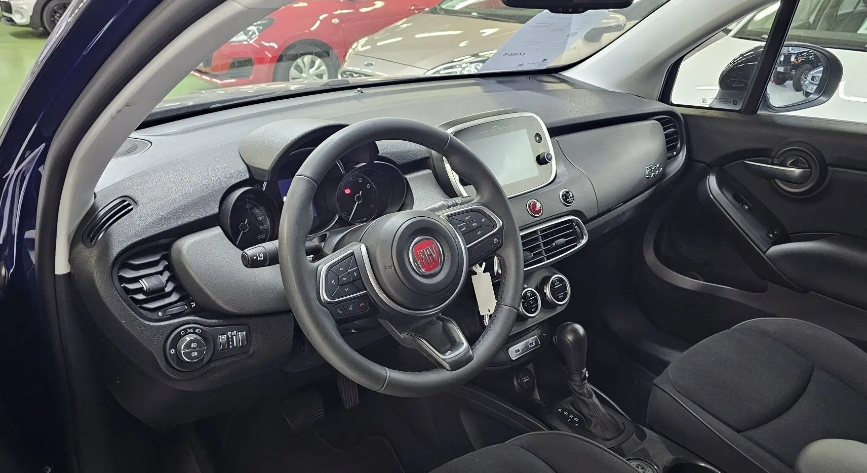 Intérieur du FIAT 500X 2024 vu côté conducteur, tableau de bord noir avec volant multifonction et écran tactile central.