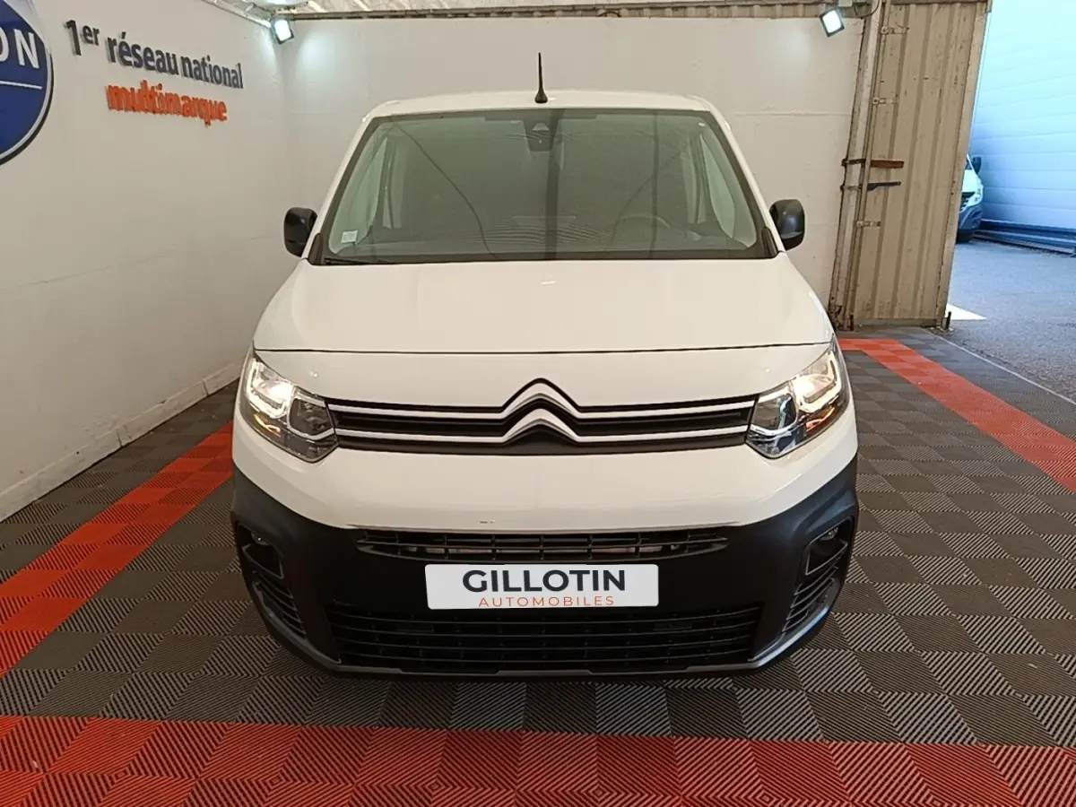 Vue frontale d’un Citroën Berlingo blanc avec pare-chocs noir et phares allumés en intérieur showroom.