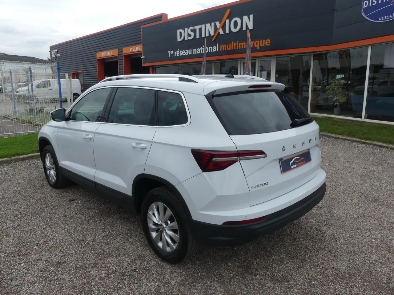 Vue 3/4 arrière droite d'un SUV SKODA KAROQ blanc garé devant un bâtiment commercial avec vitres arrière teintées.