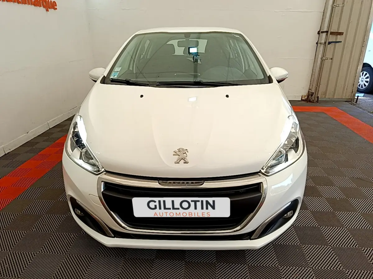 Vue avant d'une Peugeot 208 Puretech 82 Signature blanche, avec calandre chromée et phares allumés en intérieur showroom.