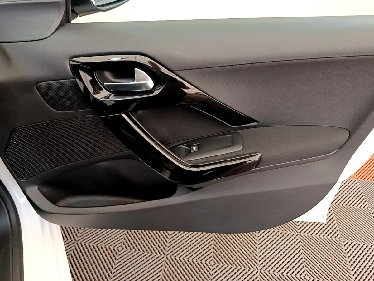 Gros plan sur la porte côté gauche de la Peugeot 208 blanche, montrant la poignée intérieure noire brillante et le bouton de lève-vitre.