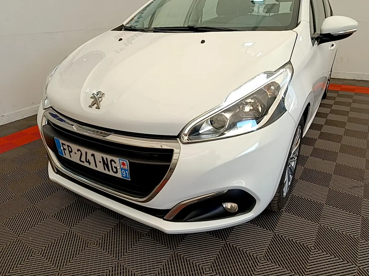 Vue 3/4 avant droite d'une Peugeot 208 blanche avec phares et calandre chromée visibles en intérieur.