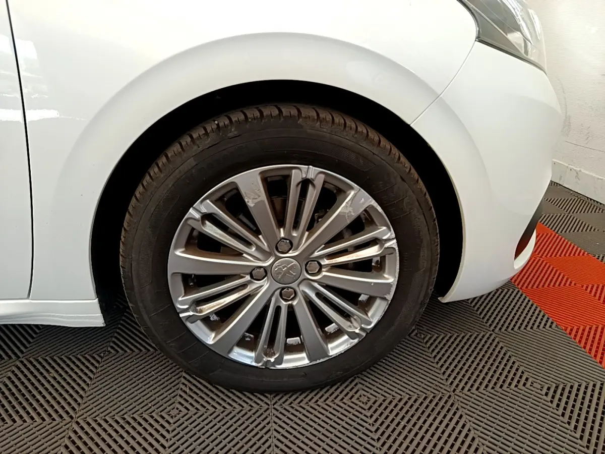 Gros plan sur la roue avant gauche d'une Peugeot 208 blanche, mettant en valeur la jante alliage au design moderne.