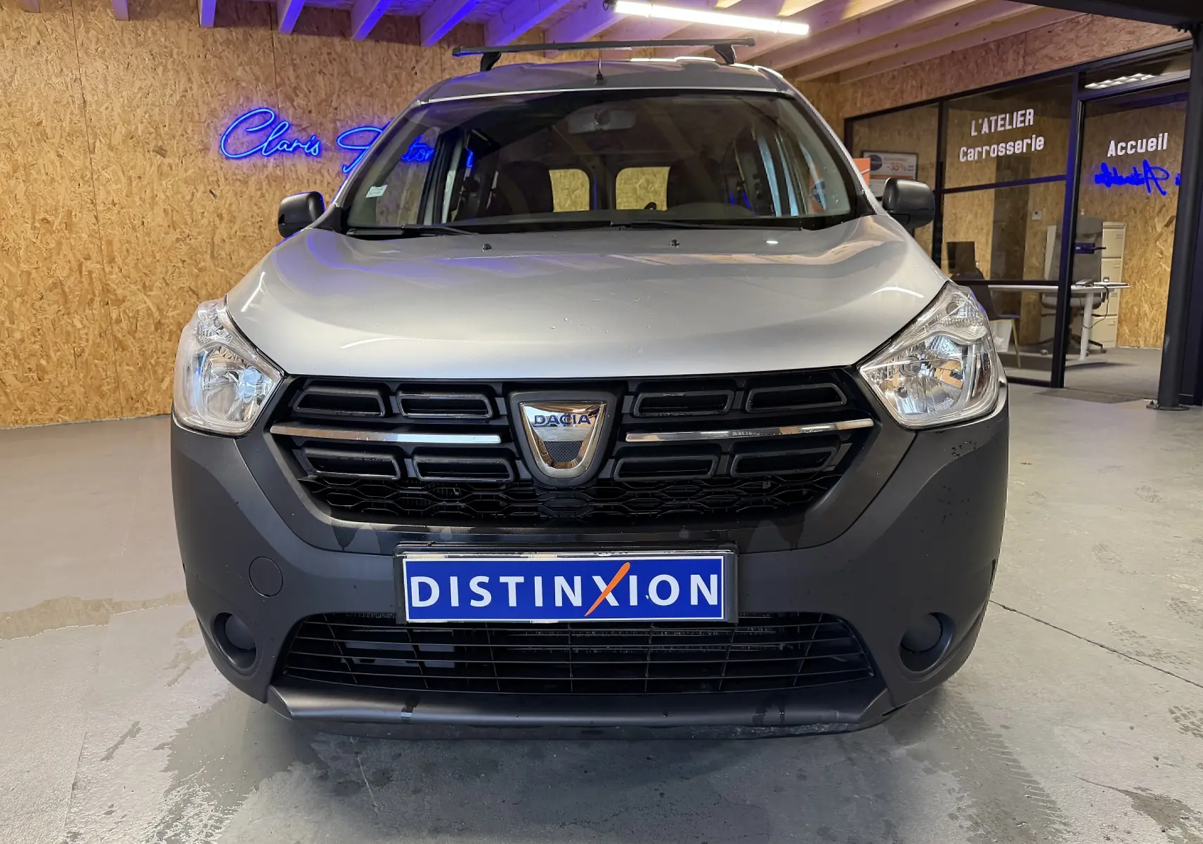 Vue frontale d'un Dacia Dokker gris avec calandre noire et logo au centre, stationné en intérieur showroom.