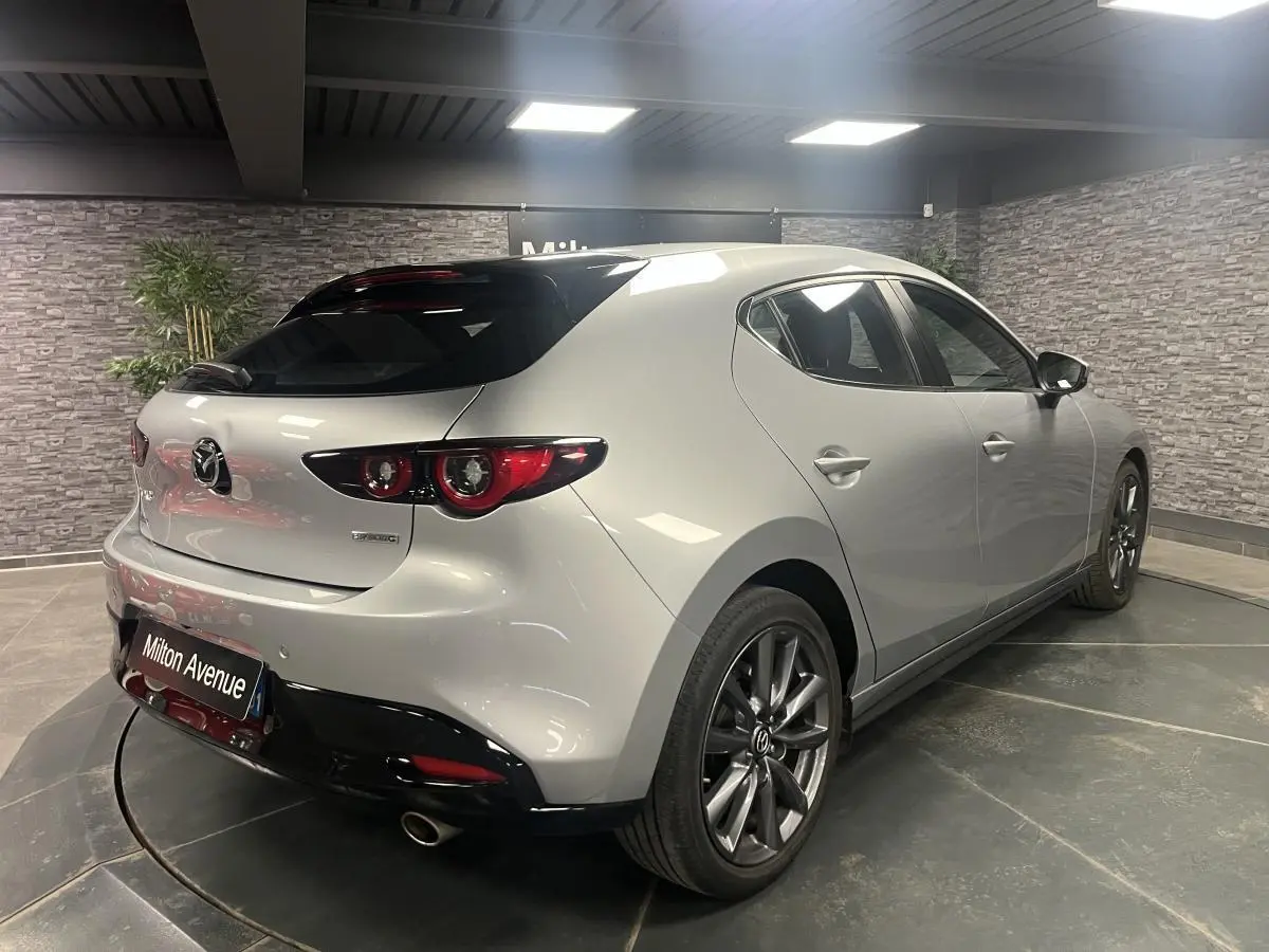 Vue 3/4 arrière droite de la Mazda3 gris métallisé avec toit noir et jantes alliage 18 pouces dans un showroom.