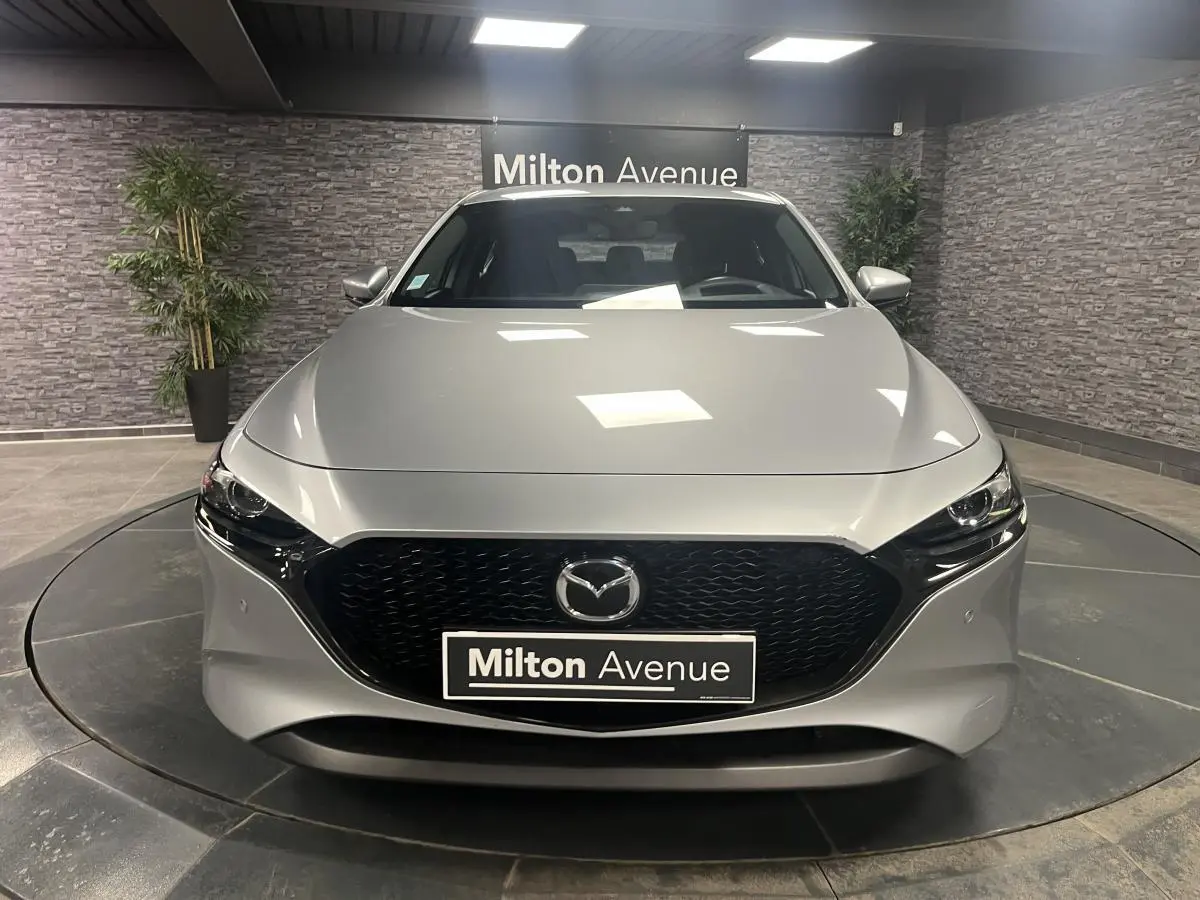 Vue frontale d'une Mazda3 gris métallisé avec calandre noire et éclairage LED, en intérieur showroom.