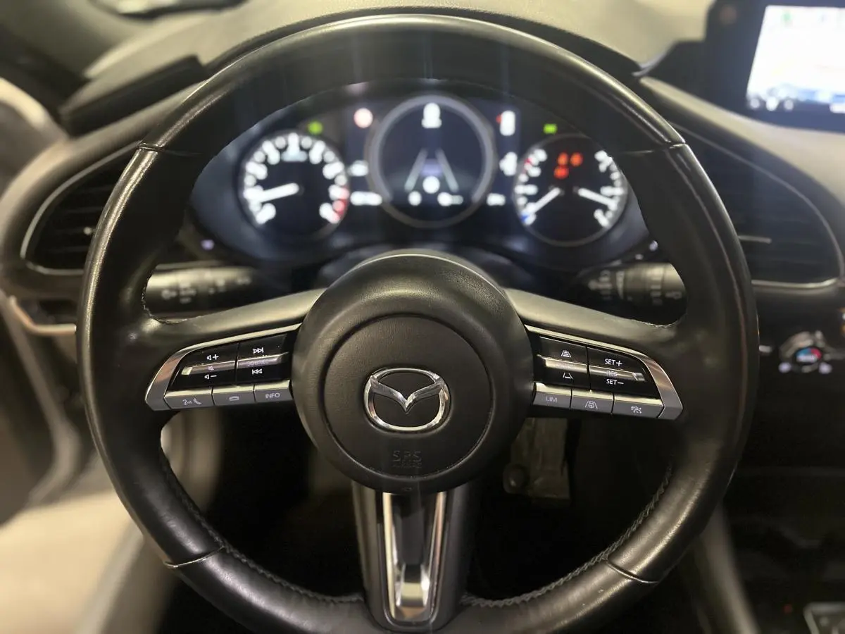 Volant cuir noir de Mazda3 2023 vu de face, avec commandes intégrées et tableau de bord numérique en arrière-plan.