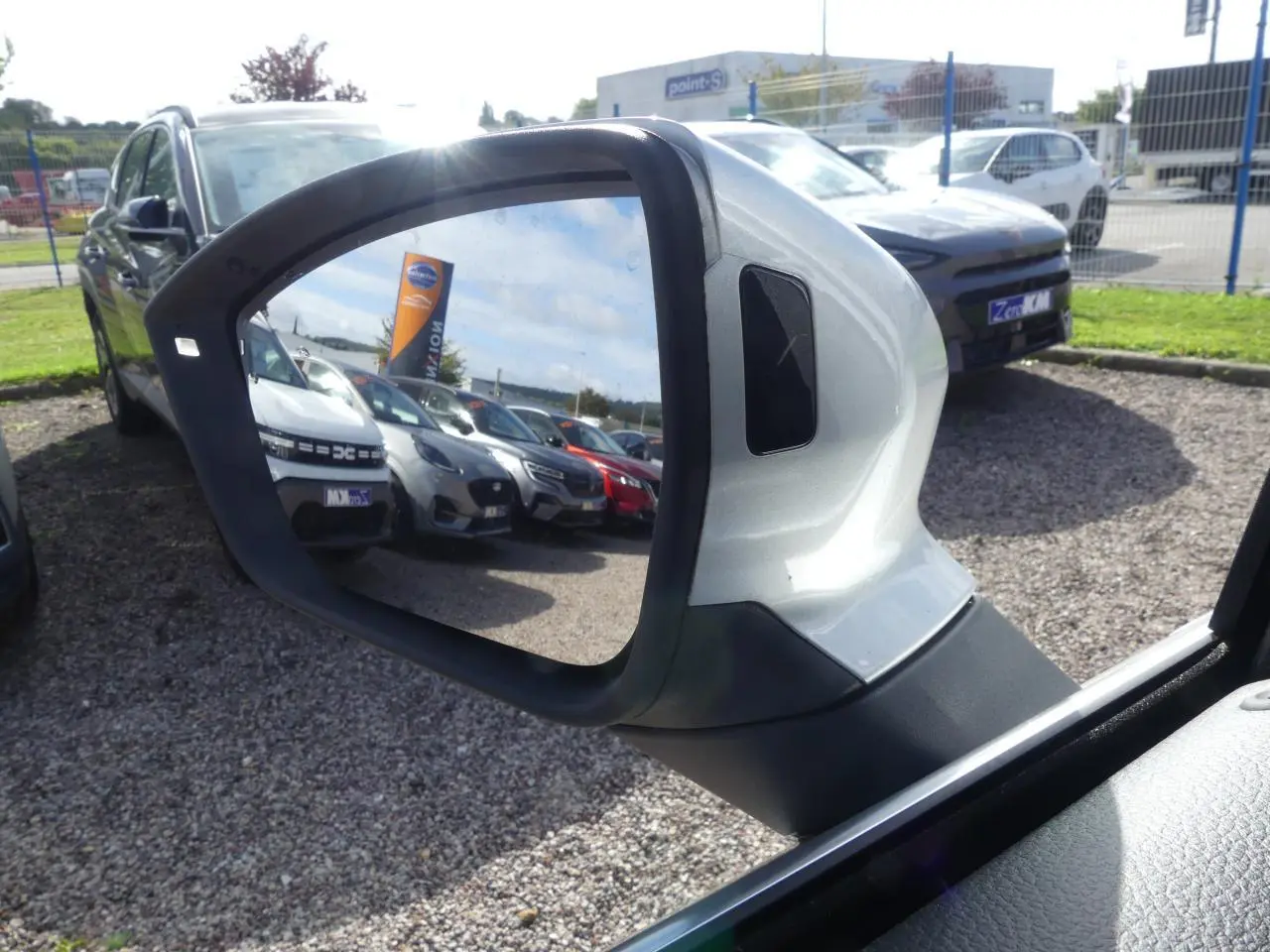 Rétroviseur extérieur droit gris clair du SEAT Ateca 2023, reflétant un parking de voitures sous un ciel partiellement nuageux.