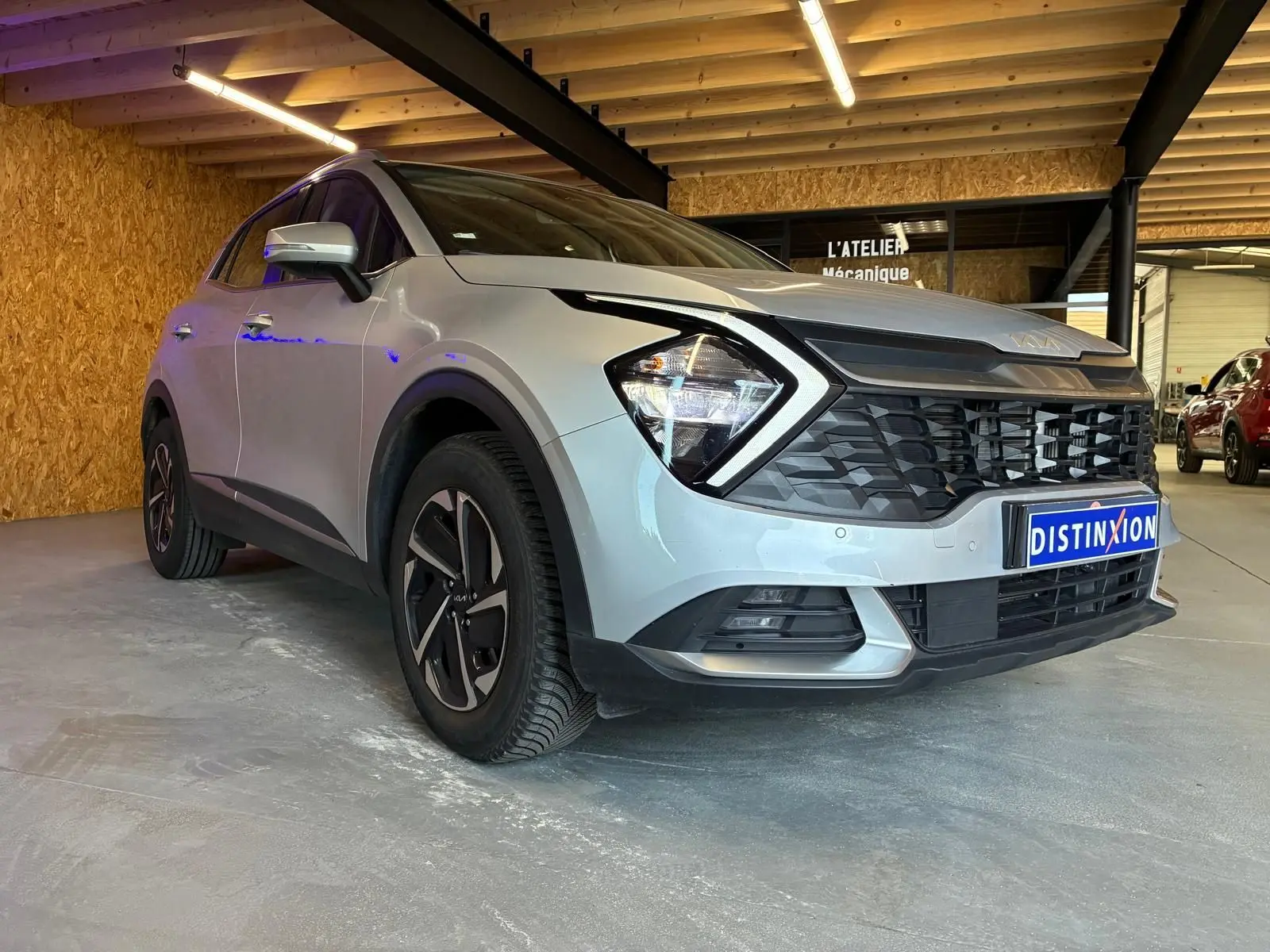 KIA Sportage hybride gris en 3/4 avant droit, avec calandre noire et phares anguleux, dans un garage éclairé.