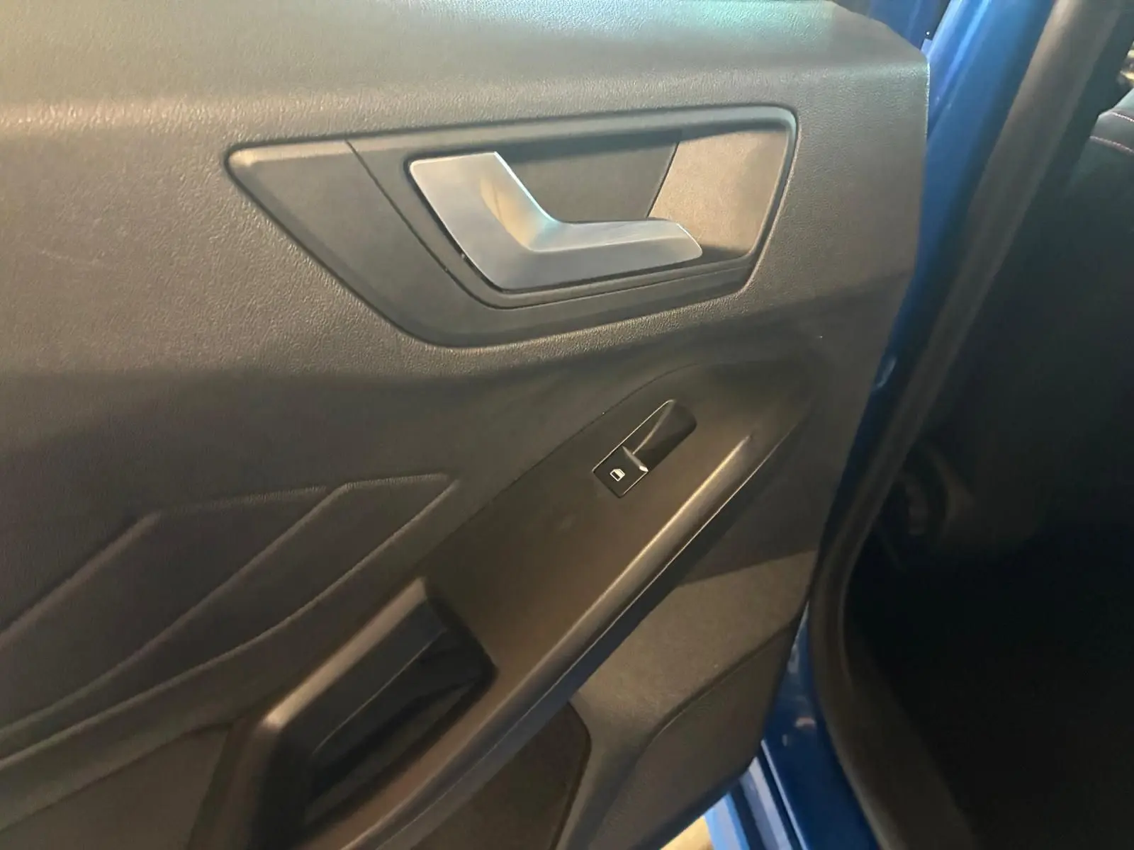 Gros plan sur la porte intérieure côté gauche d’une Ford Focus bleue 2024, avec poignée argentée et bouton de vitre électrique.