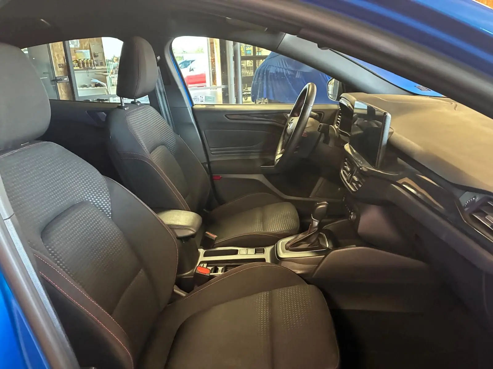 Vue intérieure côté gauche de la Ford Focus bleu 2024, montrant les sièges avant noirs et la console centrale avec boîte auto.