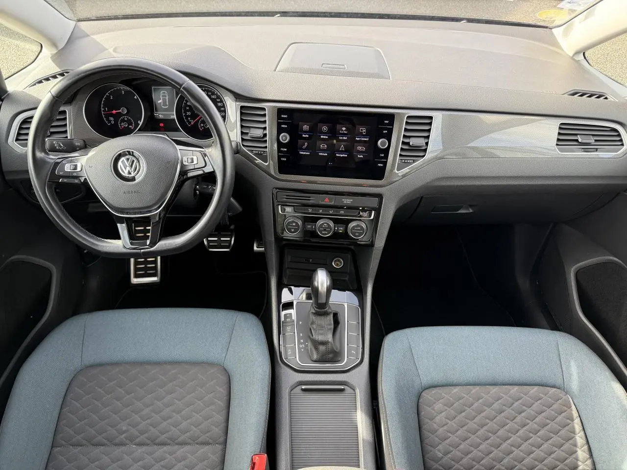 Vue intérieure avant du Volkswagen Golf Sportsvan 2019, tableau de bord noir avec écran tactile et sièges bicolores gris et bleu.