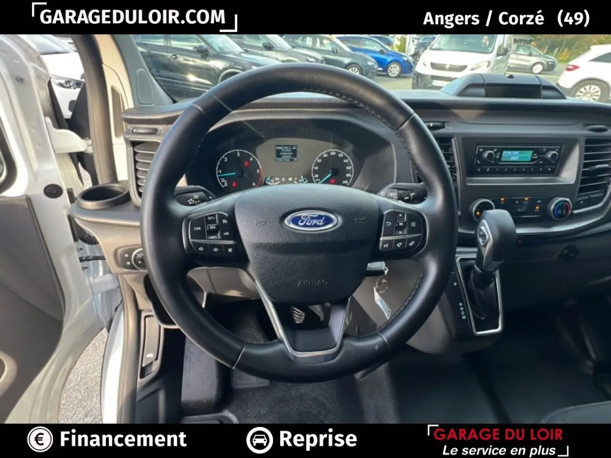 Vue intérieure centrée sur le volant cuir multifonctions d'un Ford Transit Custom blanc, tableau de bord et levier automatique visibles.