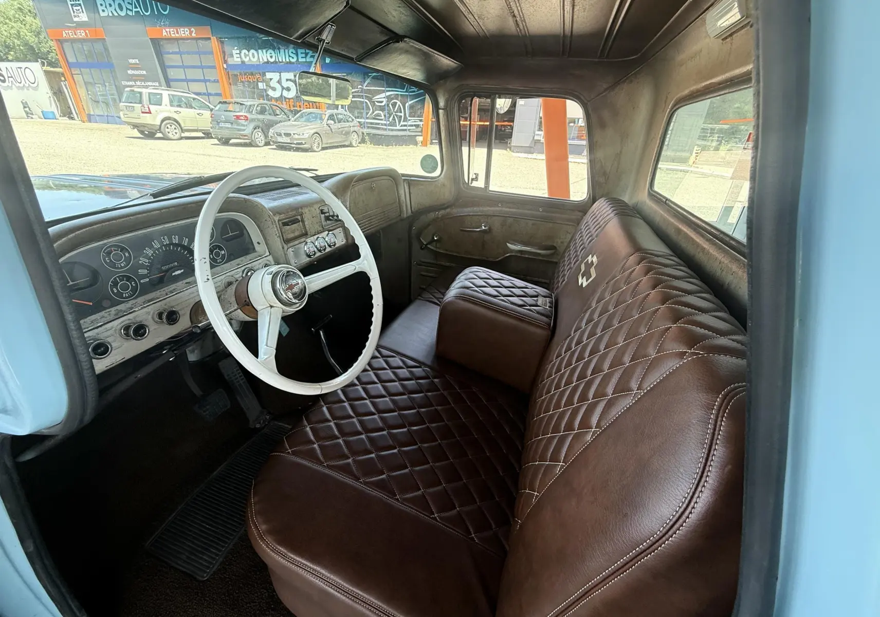 Vue intérieure côté conducteur du Chevrolet C10 Apache 1963 bleu, avec volant blanc et sièges marron matelassés.