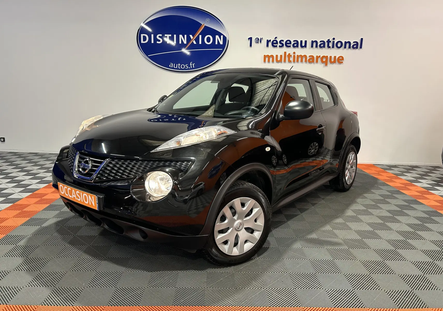 Nissan Juke noir métal en 3/4 avant droit, avec attelage visible, sur sol damier gris et orange en intérieur showroom.