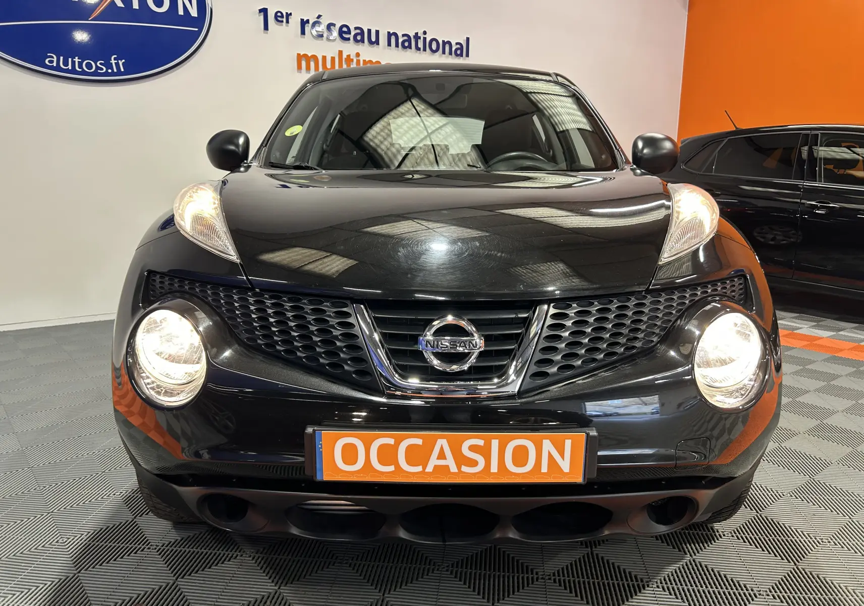 Vue avant d'un Nissan Juke noir métal avec phares allumés et plaque "OCCASION" en showroom.
