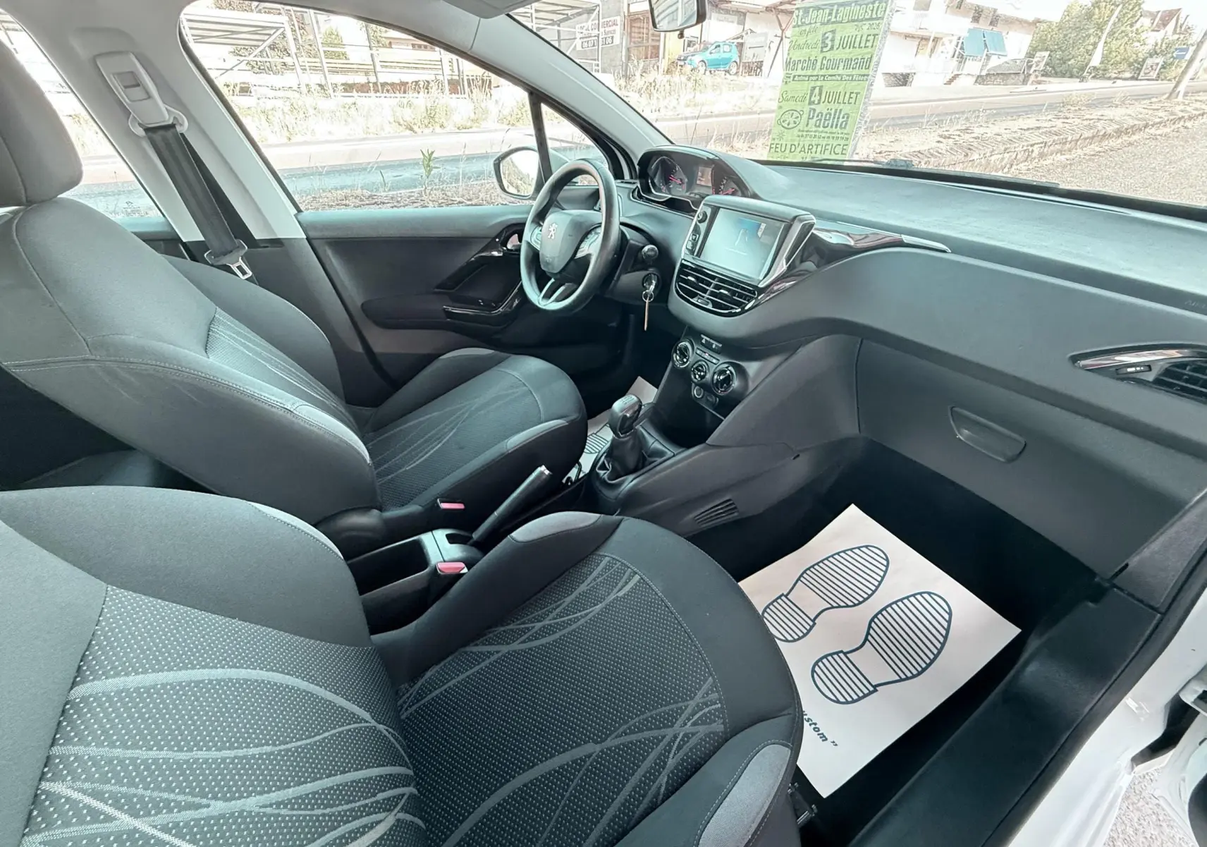 Vue intérieure côté conducteur de la Peugeot 208 blanche 2013, montrant les sièges noirs et la console centrale avec boîte manuelle.