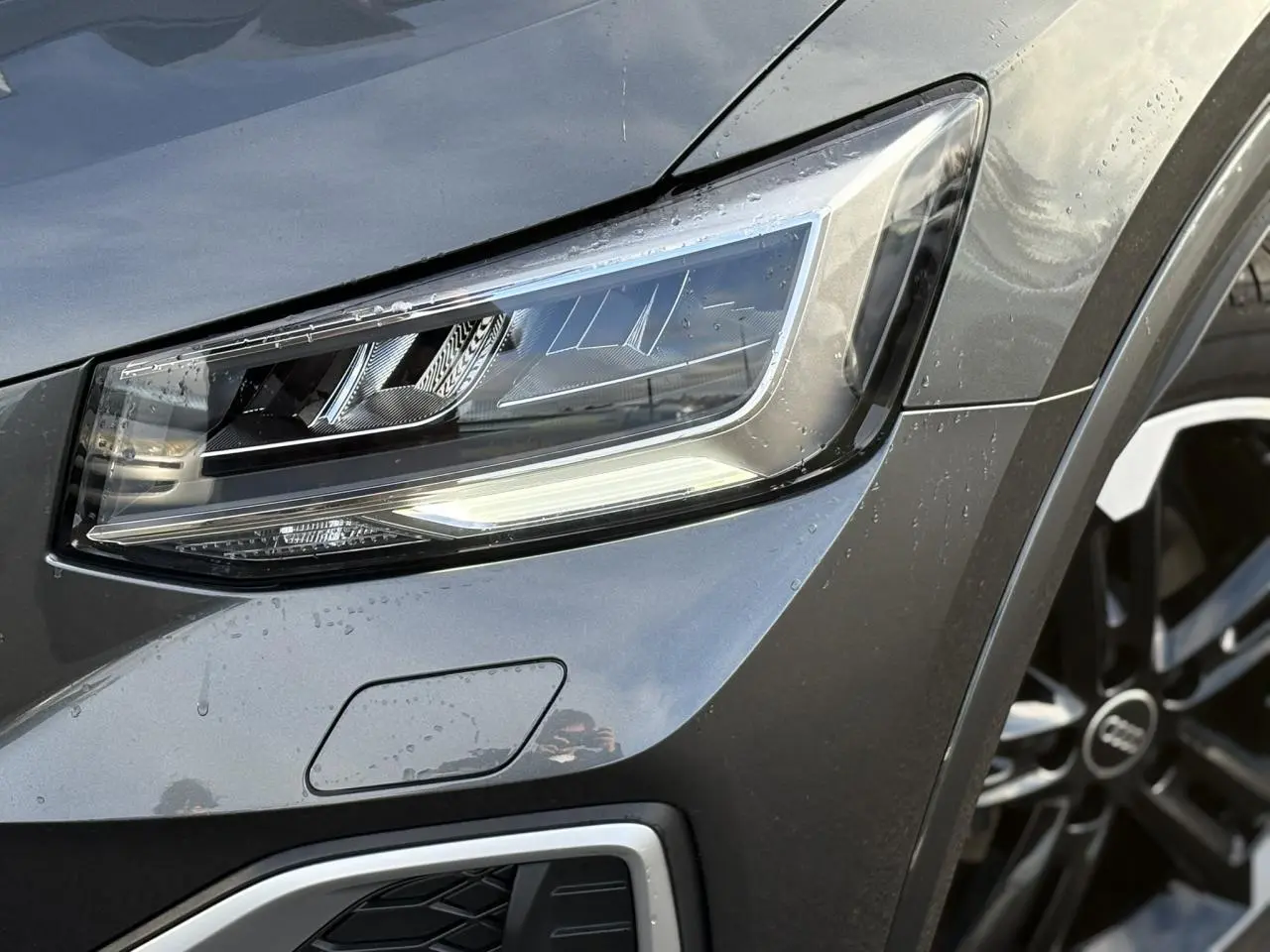 Gros plan sur le phare avant droit et la jante Audi Sport 5 branches titane mat du Q2 gris Daytona 2025.