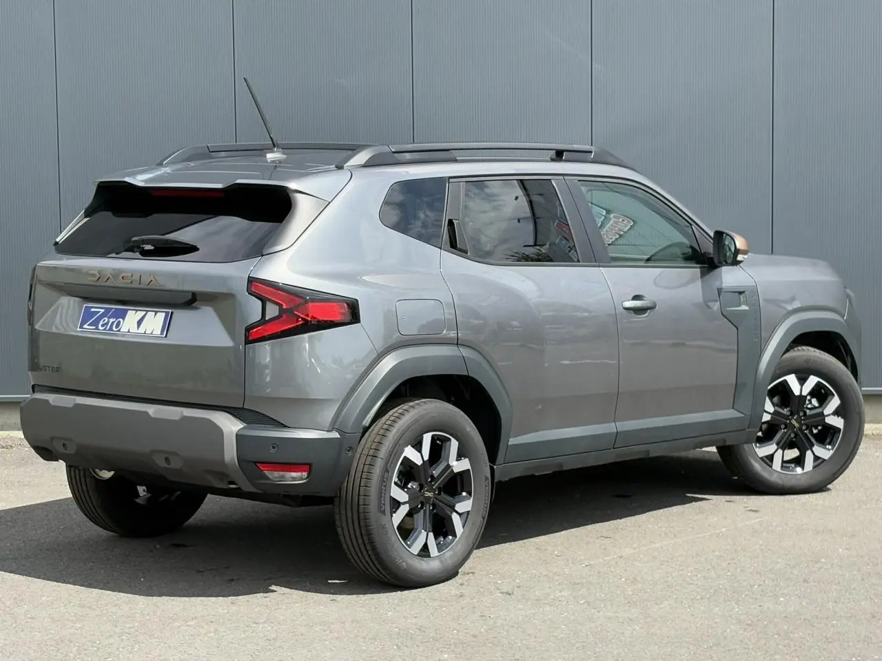 Vue 3/4 arrière droite du Dacia Duster 2025 gris schiste avec jantes alliage et barres de toit noires.