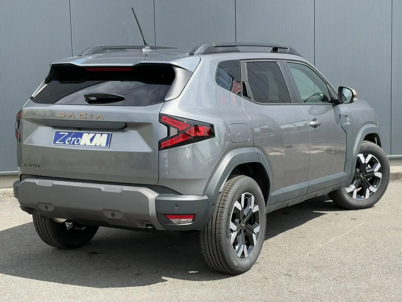 Vue 3/4 arrière droit du Dacia Duster gris schiste avec jantes alliage et barres de toit noires.