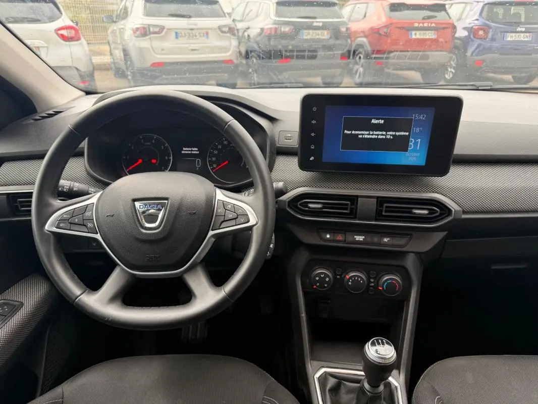 Vue intérieure du tableau de bord et volant noir d'une Dacia Sandero blanche, avec écran tactile et boîte manuelle visible.
