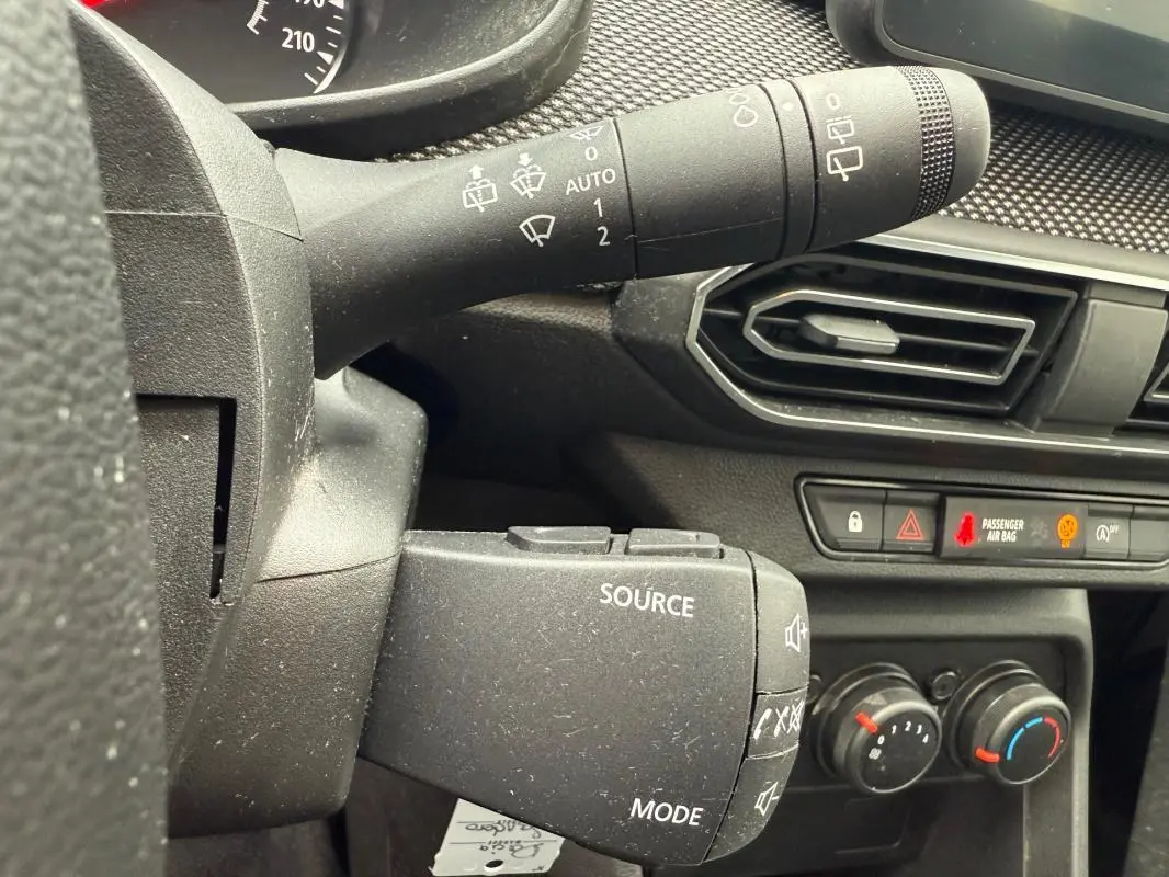 Gros plan sur la commande de source audio et les réglages de climatisation dans le tableau de bord noir d'une Dacia Sandero blanche.