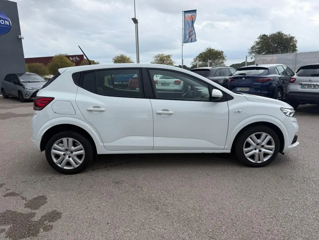 Profil droit d'une Dacia Sandero blanche 2021 avec vitres surteintées et jantes alliage en arrière-plan de parking.