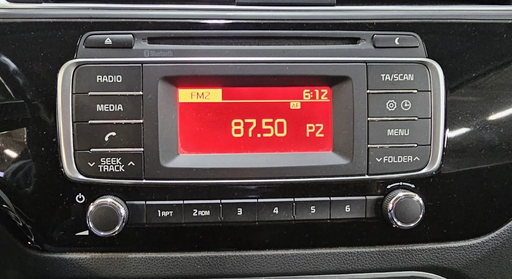 Gros plan sur la radio avec écran rouge affichant la fréquence 87.50 FM dans le tableau de bord noir d'une Kia Rio III 2015.