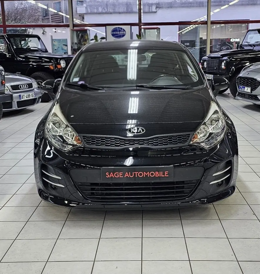 Vue frontale d'une KIA RIO III noire de 2015, exposée en intérieur avec plaque "SAGE AUTOMOBILE" visible.