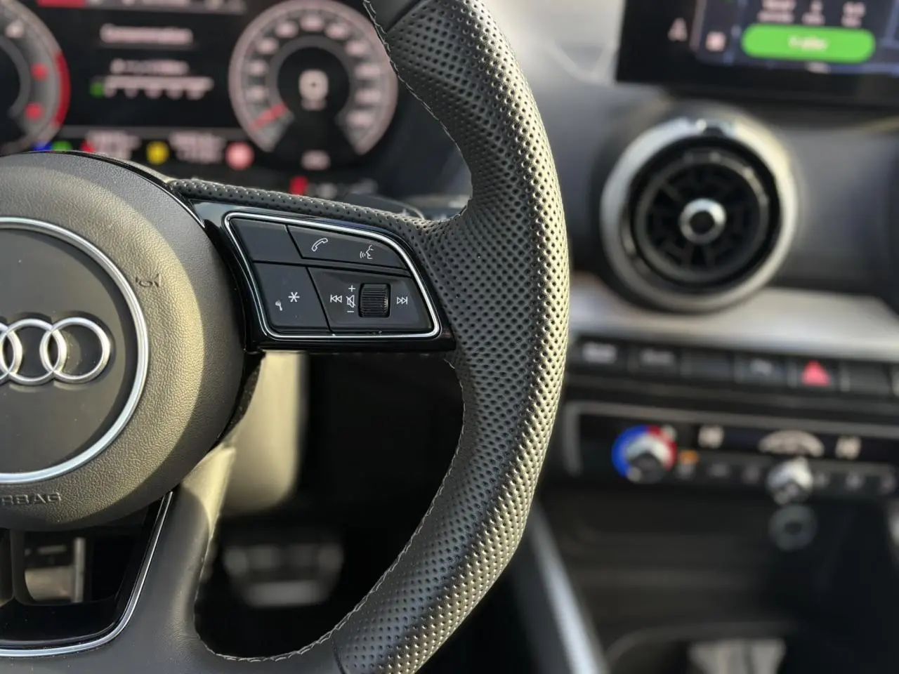 Gros plan sur le volant cuir perforé de l'Audi Q2 35 TDI S-Line avec tableau de bord numérique flou en arrière-plan.