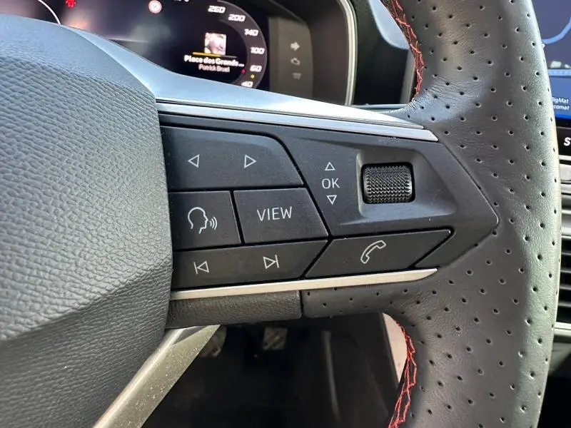 Gros plan sur les commandes gauche du volant perforé noir du SEAT LEON ST 2025, avec touches multifonctions et molette.