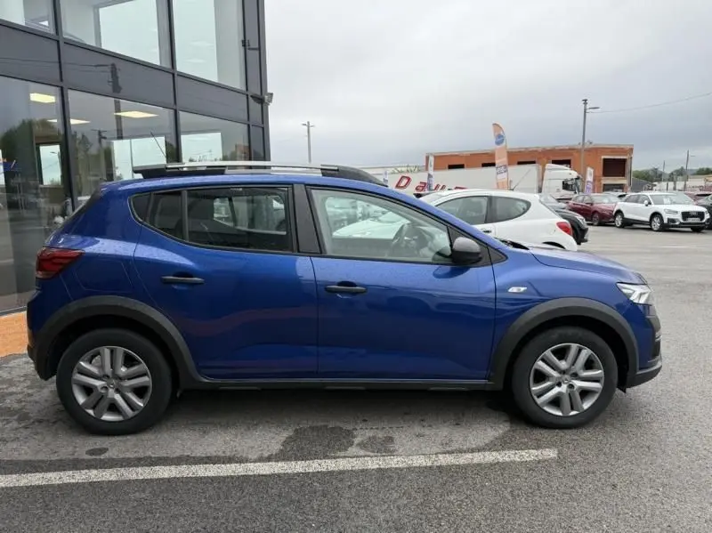 Profil côté gauche d'une DACIA SANDERO STEPWAY bleu métal 2022 avec barres de toit et protections noires sur les passages de roues.
