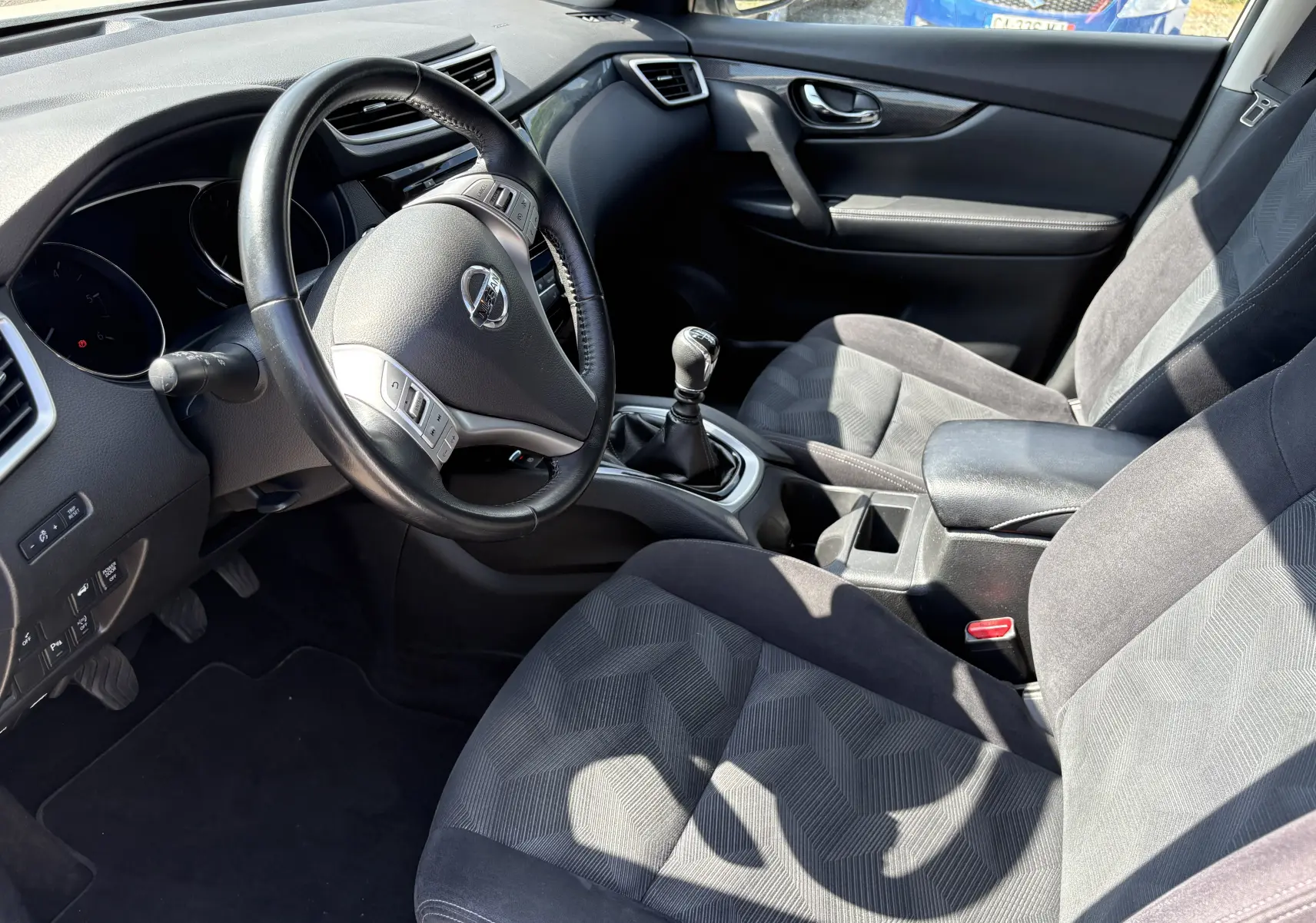 Vue intérieure côté conducteur du Nissan X-TRAIL 2016, volant cuir, sièges tissu gris et levier de vitesse manuel.