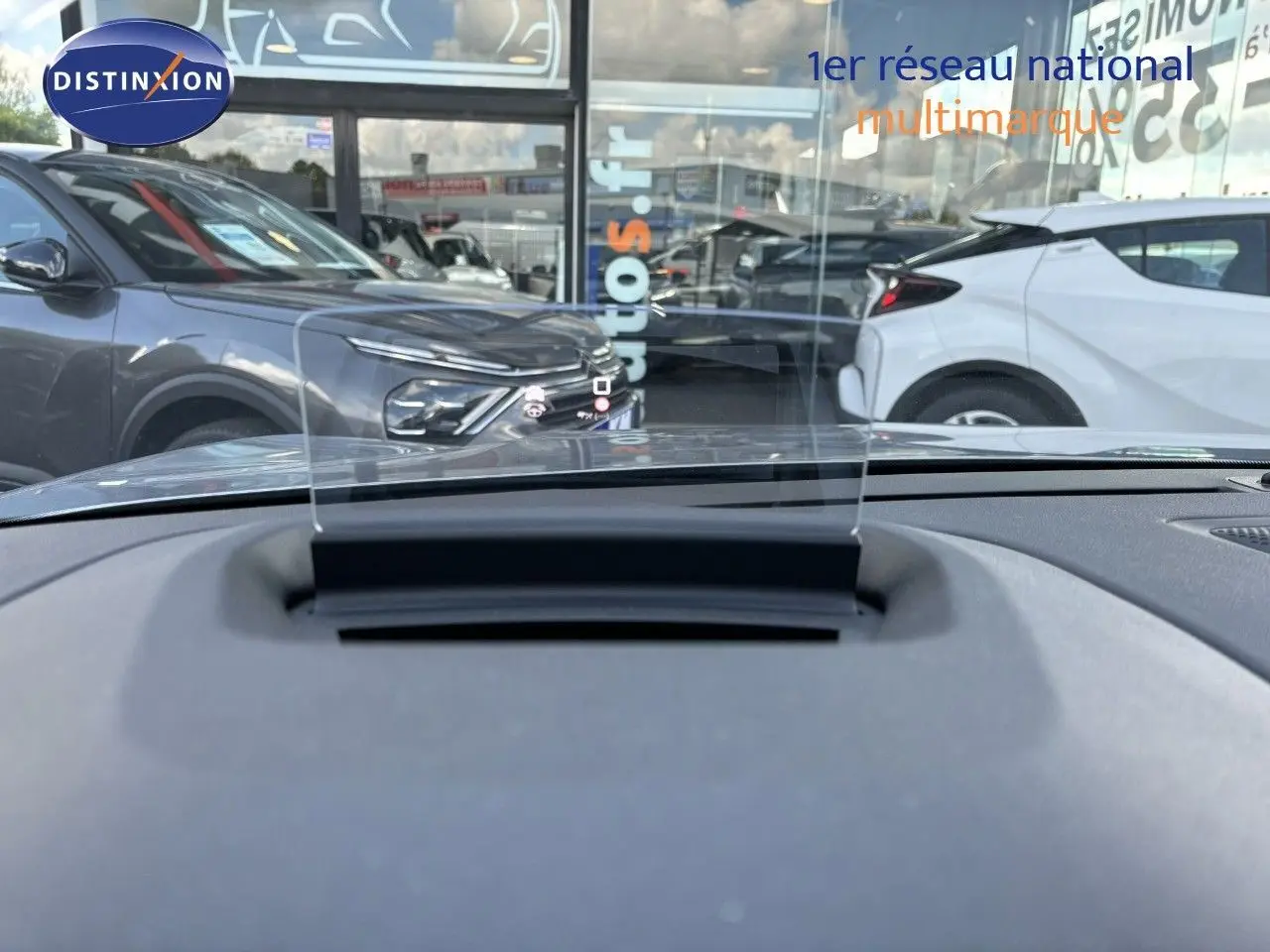 Vue depuis l'intérieur sur le tableau de bord avec affichage tête haute, devant une Citroën C4 gris Artense en 3/4 avant gauche.