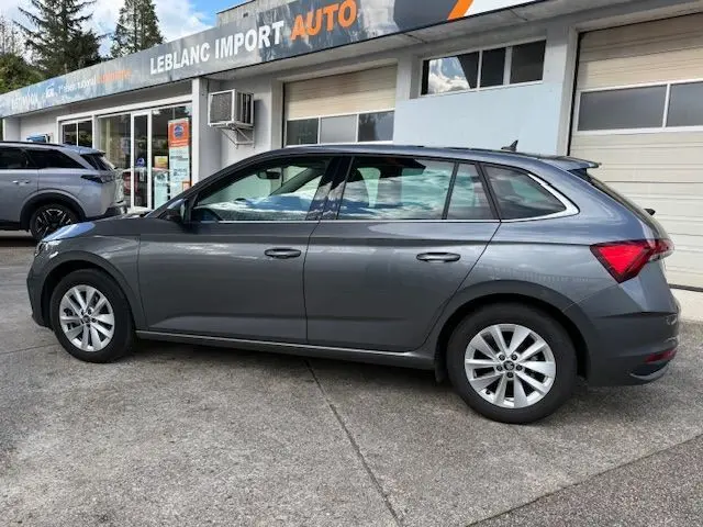 Vue de profil côté gauche d'une Skoda Scala gris graphite 2025 garée devant un garage avec jantes alliage et vitres teintées.