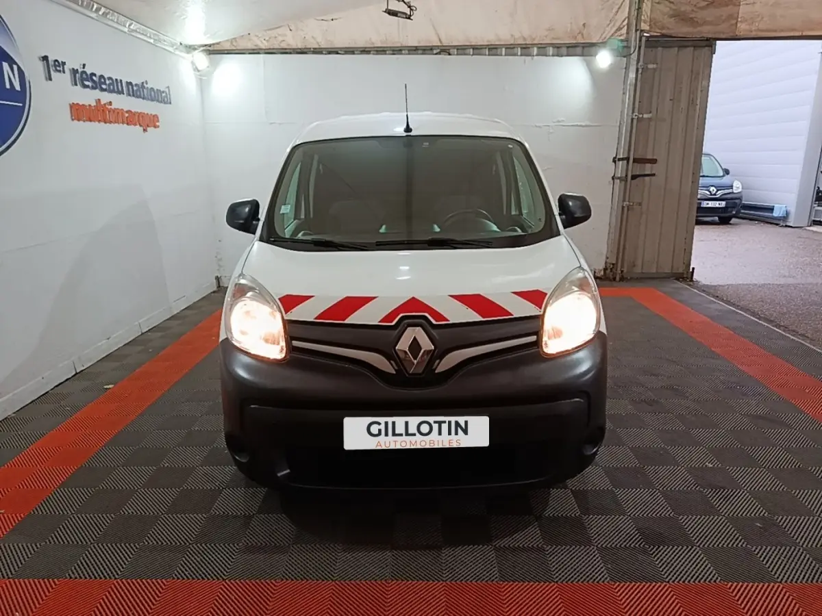 Vue avant d’un Renault Kangoo blanc 2020 avec bandes rouges diagonales sur le capot et phares allumés dans un garage.