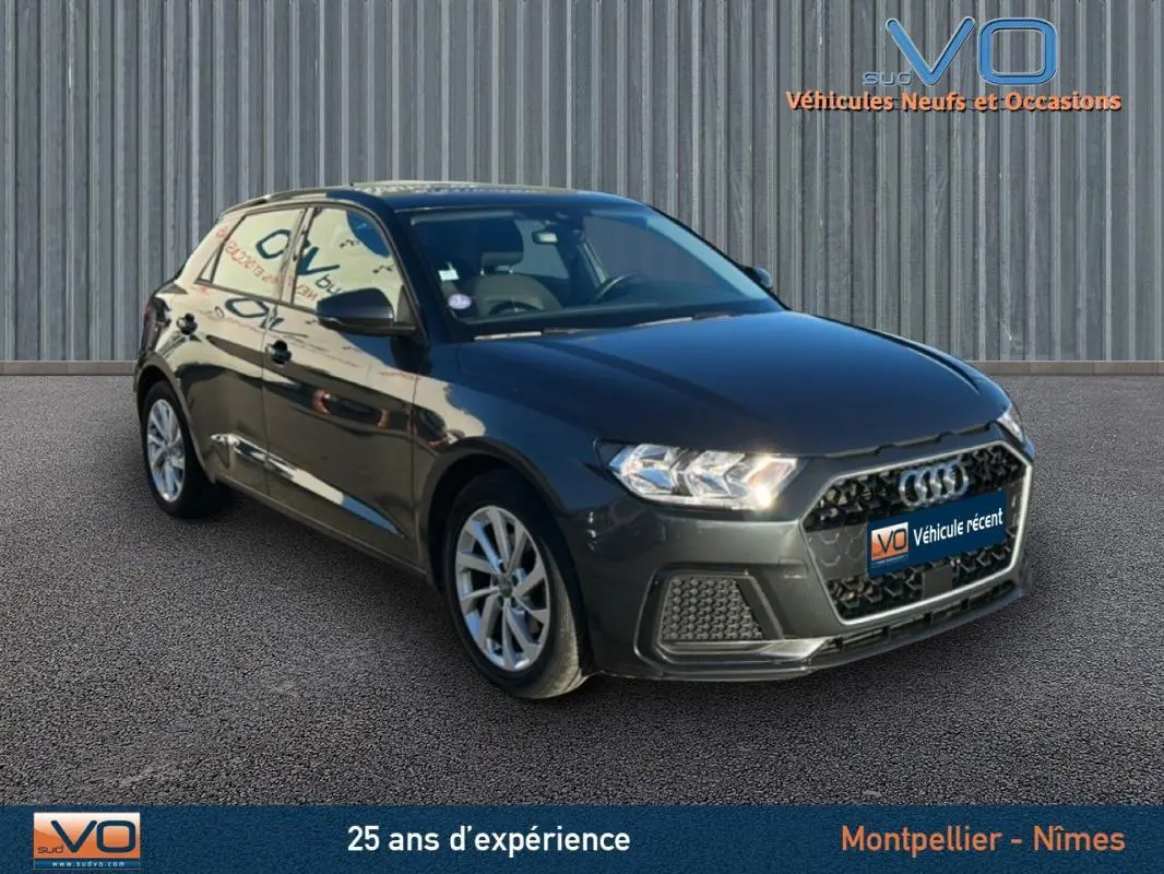 Audi A1 Sportback gris foncé vue 3/4 avant droit, avec phares allumés et jantes 16 pouces sur sol bitumé.