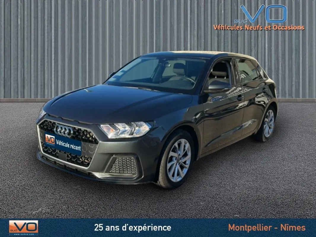 Audi A1 Sportback gris foncé vue 3/4 avant droit sur sol bitumé avec arrière-plan métallique vertical.