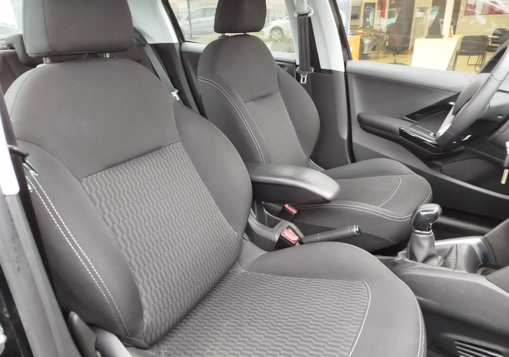 Vue intérieure côté conducteur de la Peugeot 208 2019, sièges tissu noir avec coutures blanches et levier de vitesse manuel.