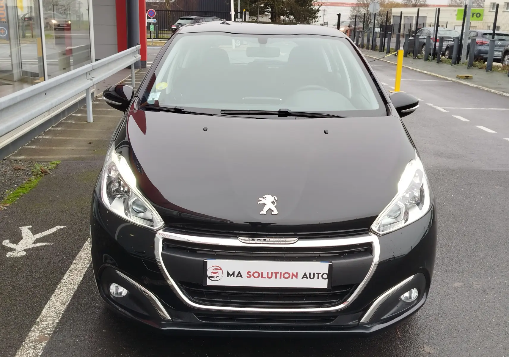 Vue avant d'une Peugeot 208 noire Perla Nera de 2019 avec phares allumés et calandre chromée visible.