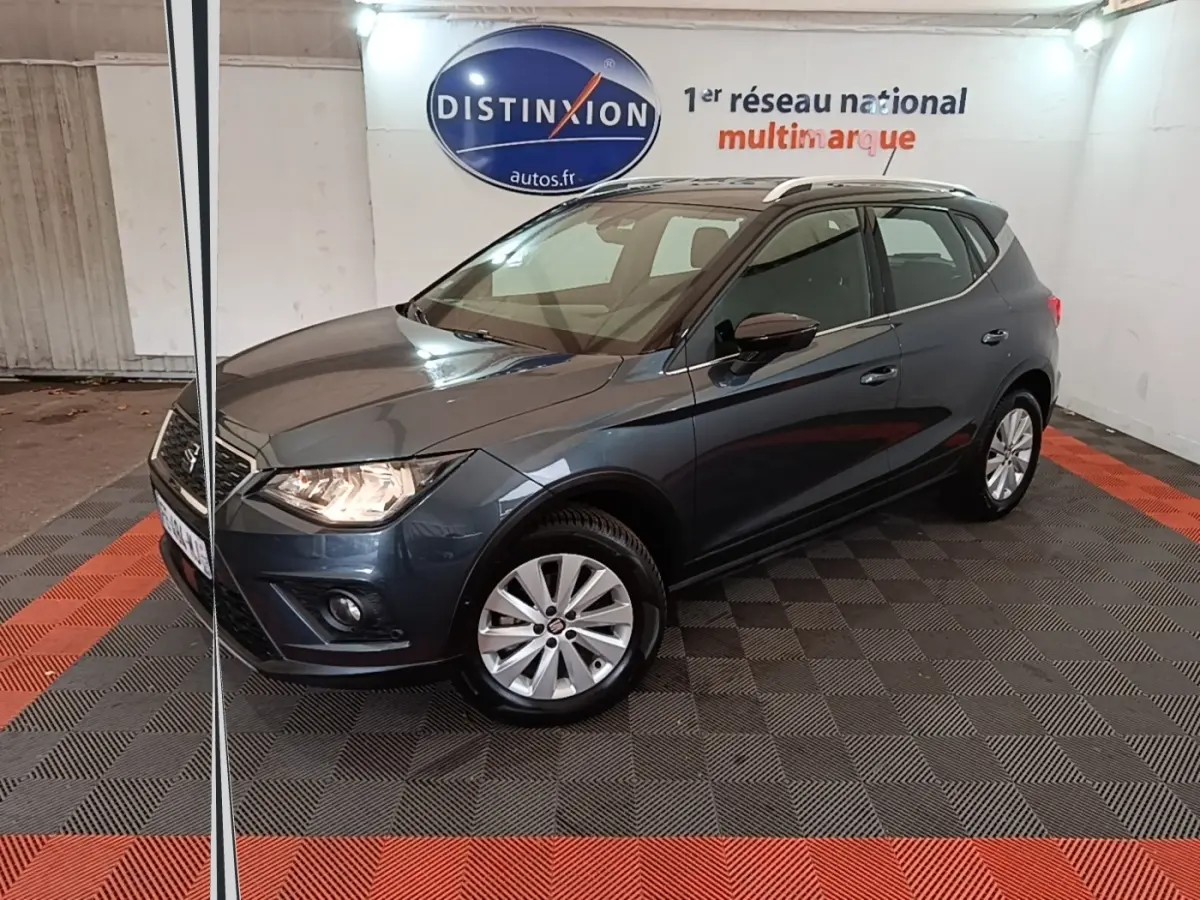 SEAT Arona gris clair en 3/4 avant droit, avec jantes alliage et feux allumés dans un showroom intérieur.
