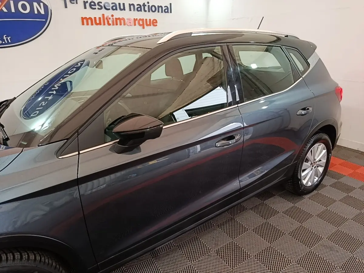 Vue latérale droite d'un SEAT Arona gris clair 2019, avec rétroviseur noir et jantes argentées, en intérieur showroom.