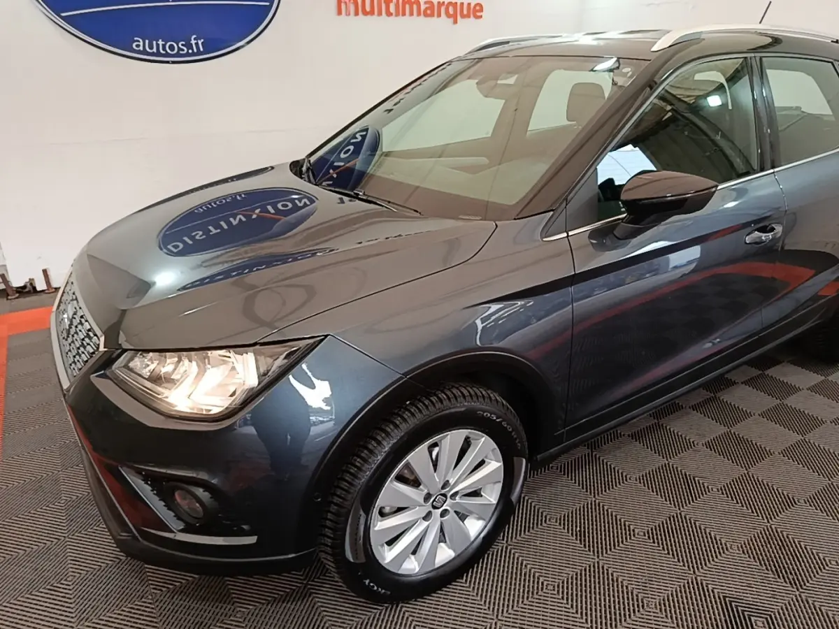 SEAT Arona gris clair vue 3/4 avant droit, phares allumés et jantes alliage visibles en intérieur showroom.